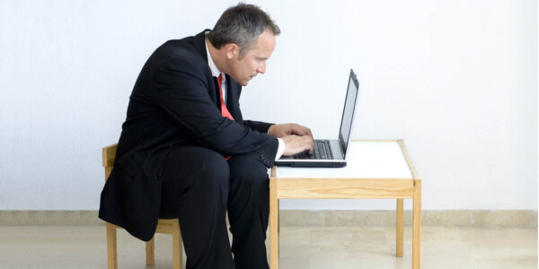 A businessman sits at a child size table and chair working on his laptop.